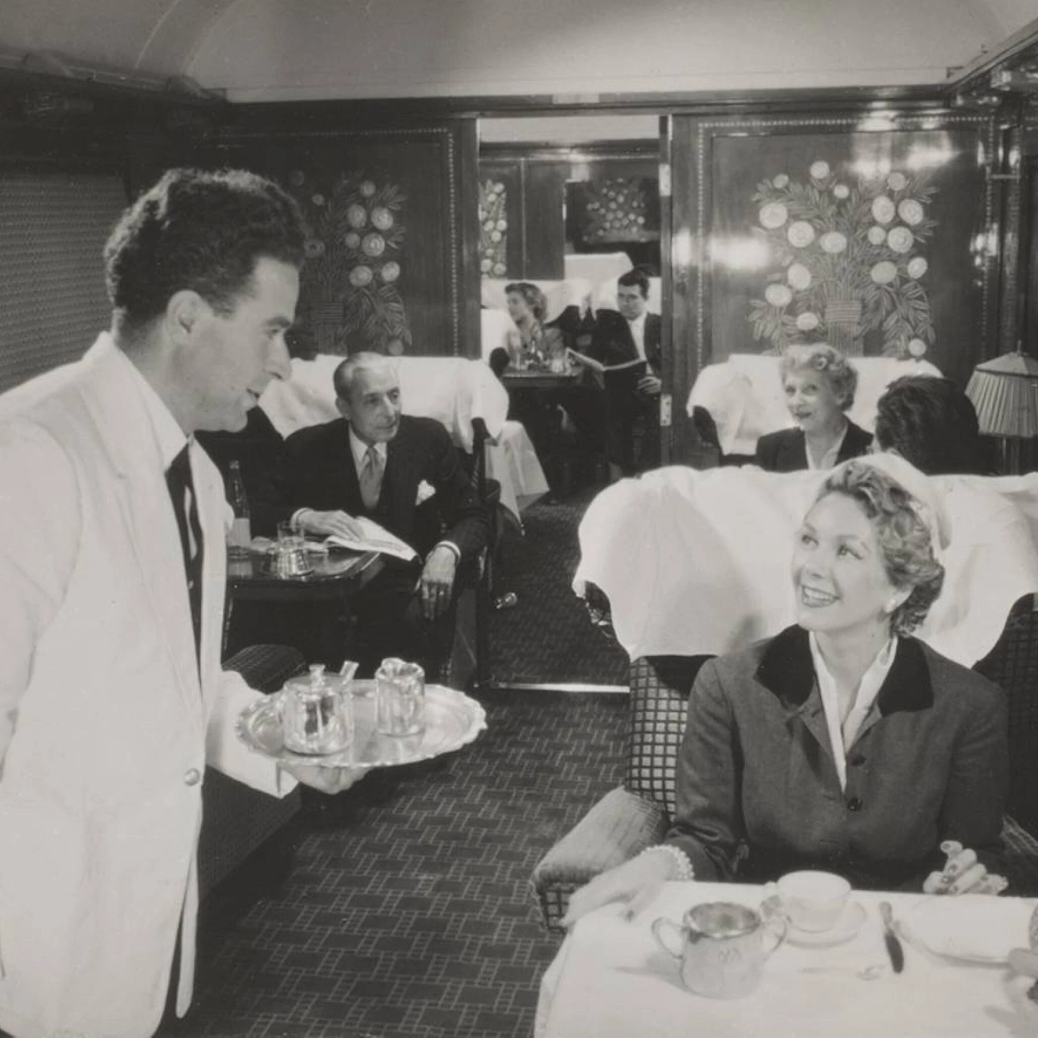 Orient Express waiter serving tea to a smiling passenger in a 1926-style dining carriage with decorative Lalique panels 