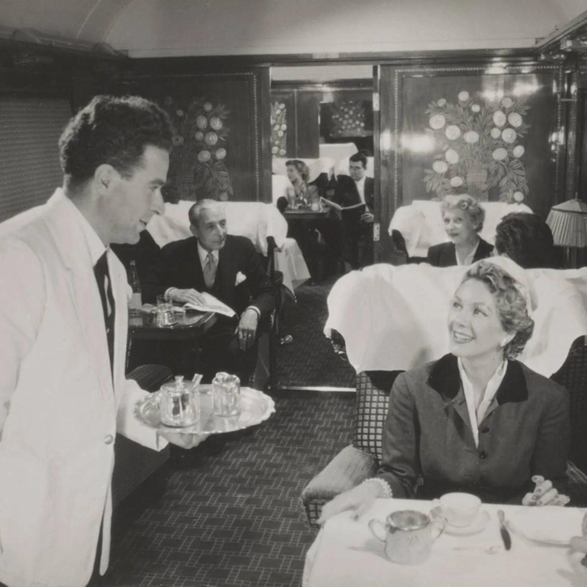 Orient Express waiter serving tea to a smiling passenger in a 1926-style dining carriage with decorative Lalique panels