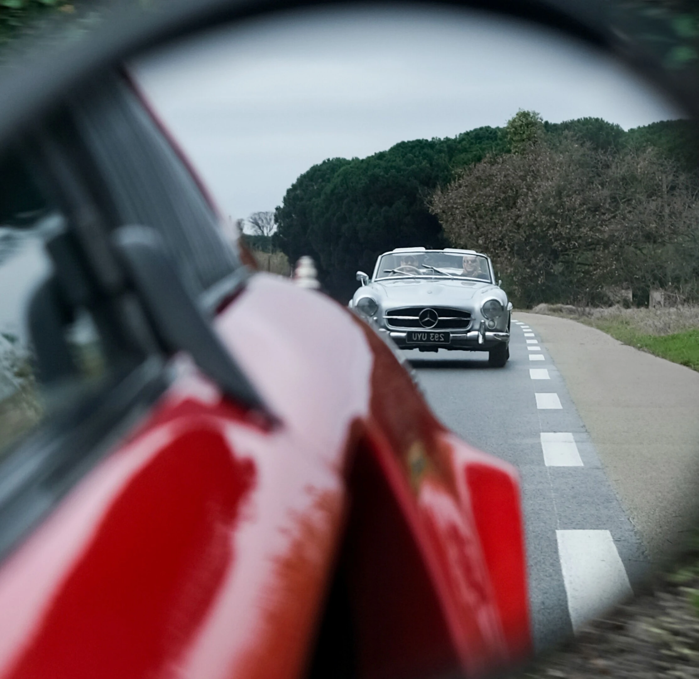 Saint-Tropez car rally: silver Mercedes 300SL convertible driving on a narrow country road behind a red classic car 